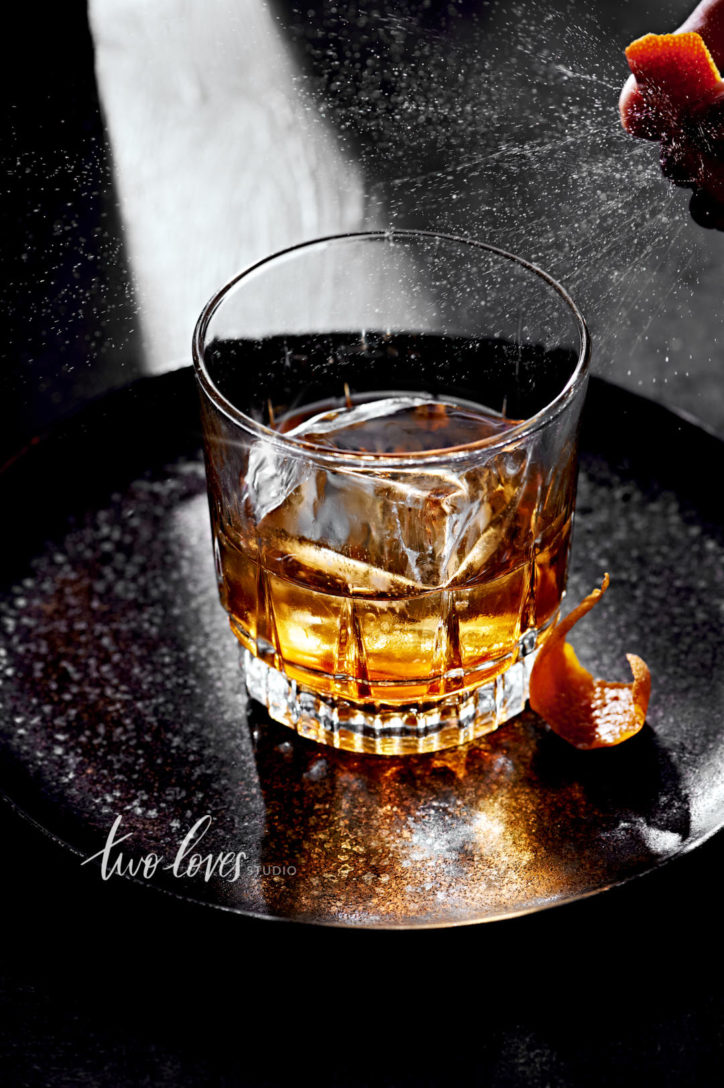 Drink Photo of a whiskey in a rocks glass on a black plate with an orange garnish. 