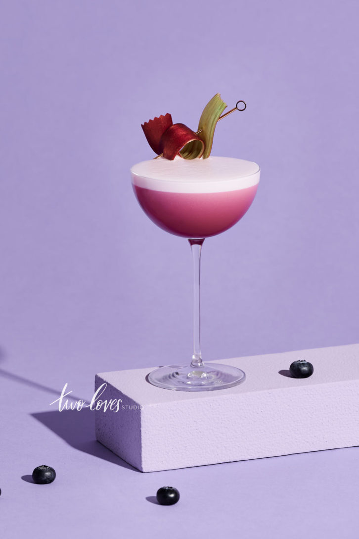 A stunning pink cocktail with a thick dry shake foam on top with a purple backdrop, resting on a stand with berries around it and a fancy garnish.