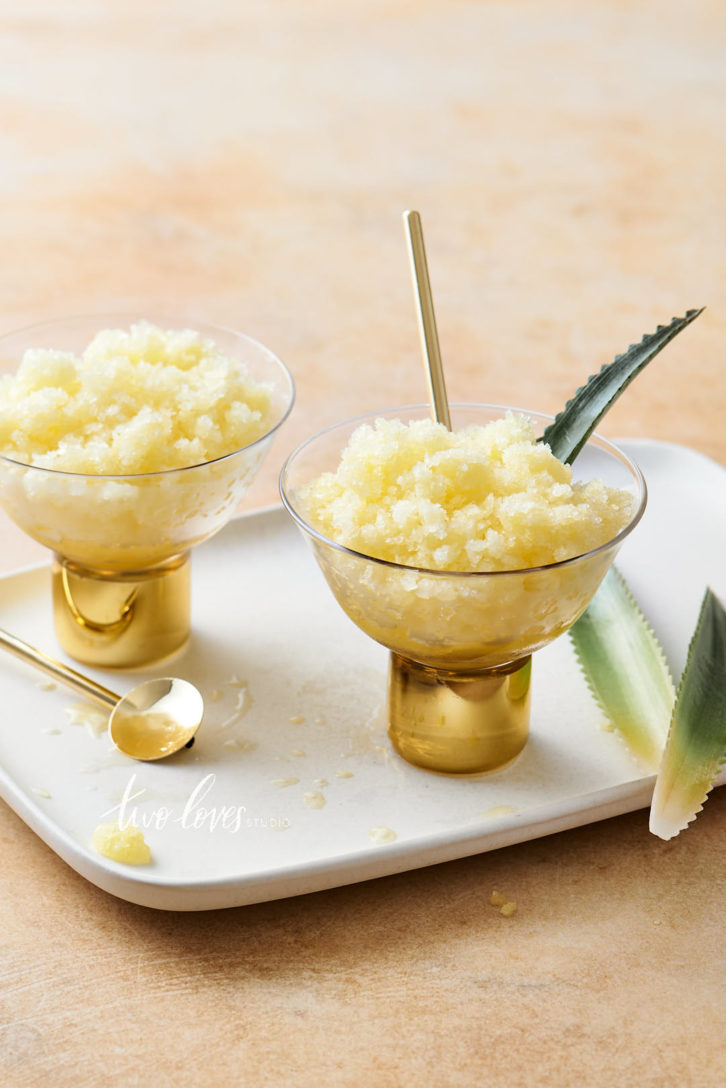 Pineapple leaves on a side dish with one in a golden frozen cocktail on a peach coloured backdrop.  