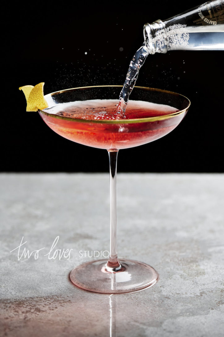 Cosmopolitan cocktail with a lemon wedge and sparking water being poured in from the top right. 