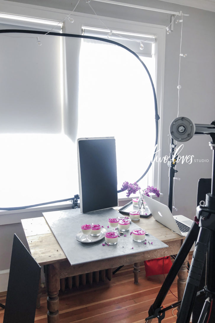 Wide view example set up shot of equipment for beginner food photographers, including a reflector, tripod, wooden table with desserts on-top.