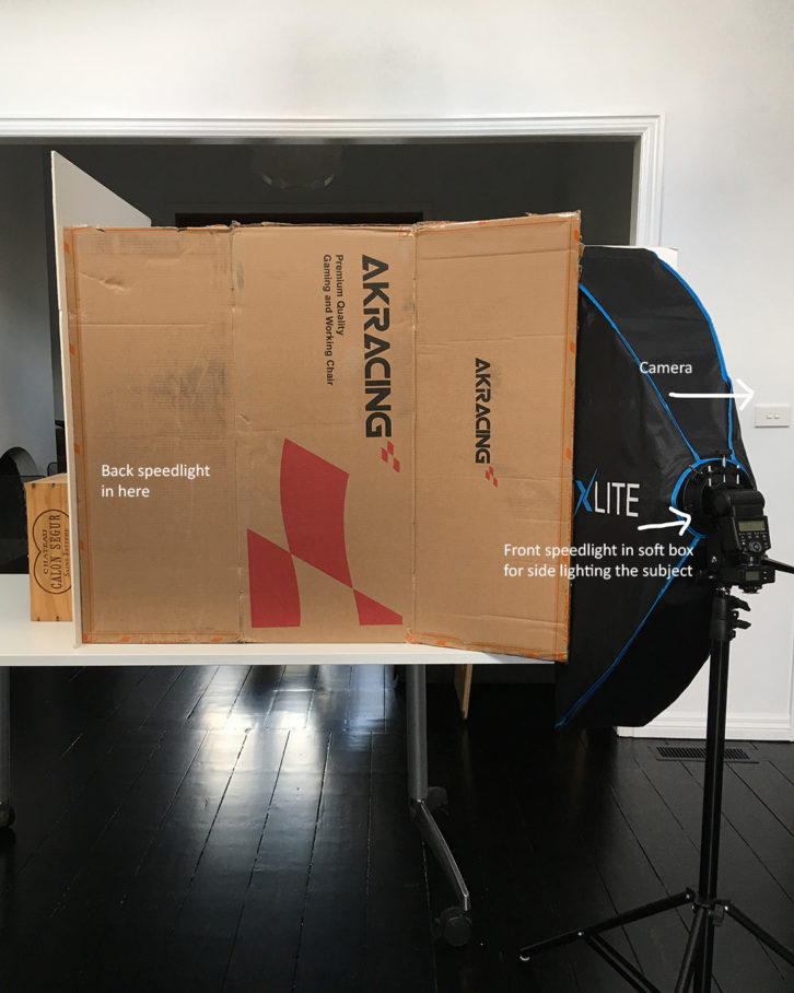 An example of flash for product photography. Showing a white table with a large cardboard box on top. A large white foam card behind the cardboard box. A front speedlight in soft box. 