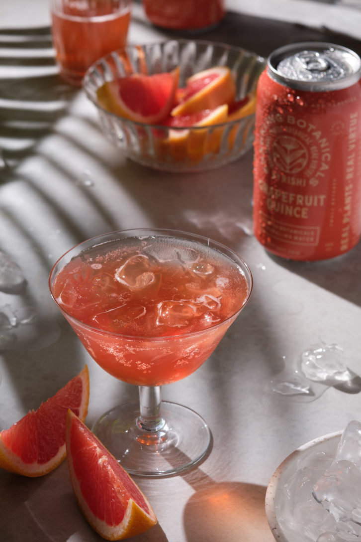 A marble background with a fern casting dappled light on a sliced orange and beverage.