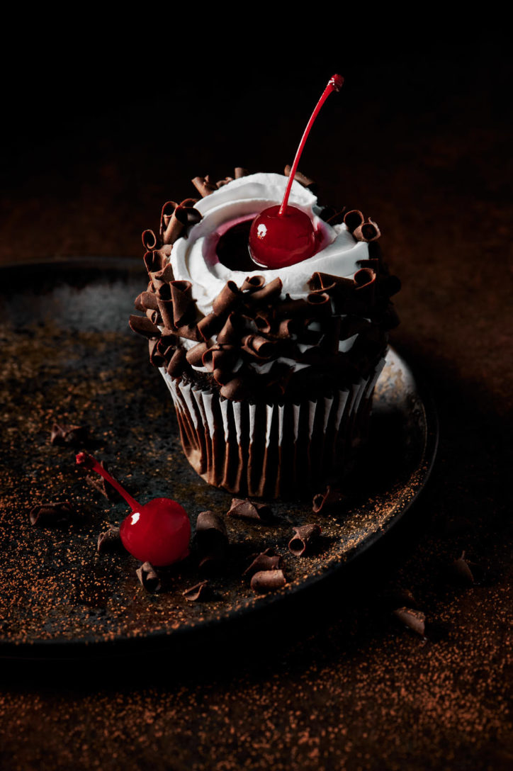 Chocolate cupcake with white icing and a cherry on top of a black plate and chocolate dusting. 