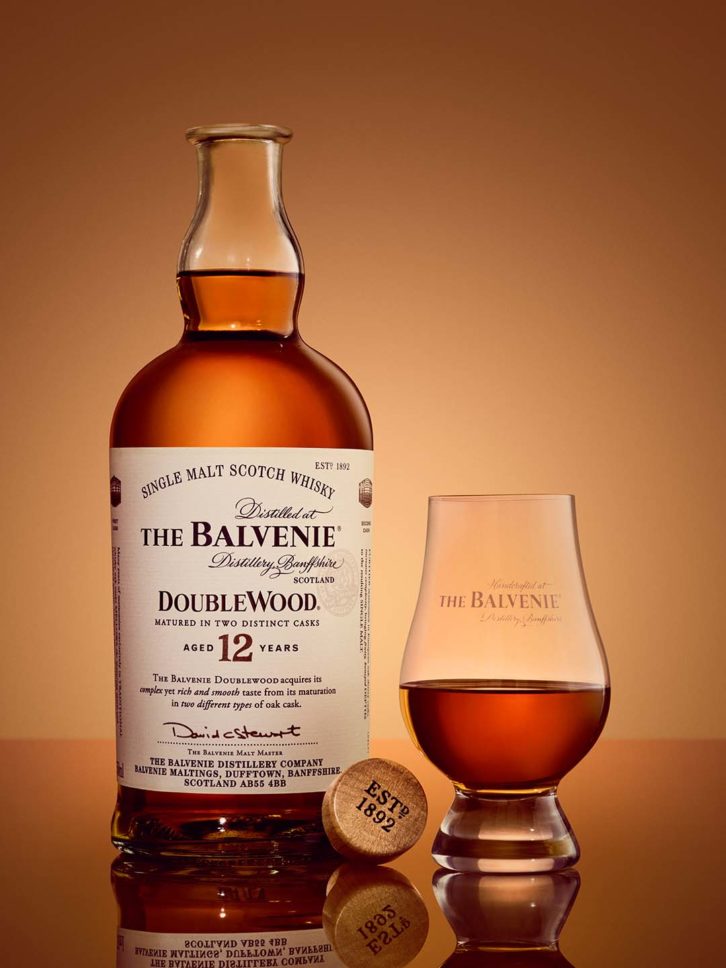 A bottle of Balvenie Doublewood Scotch whisky, with a small glass next to it on a reflective surface. 
