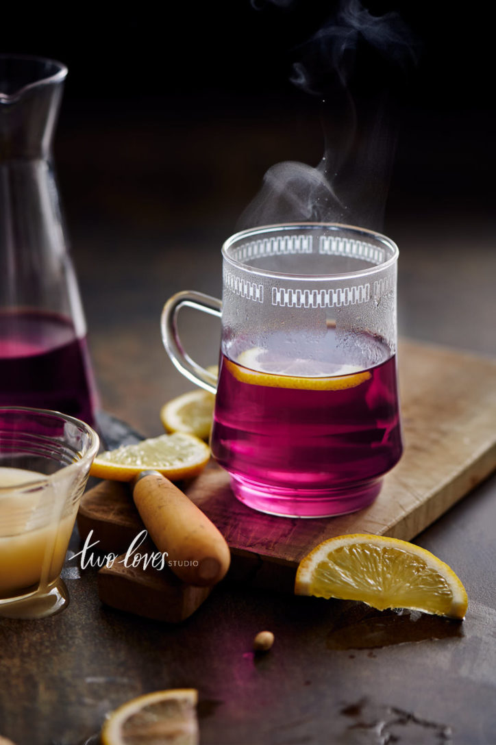Tall glass teacup with a pink, purple liquid and slices of lemon on a wooden cutting board.