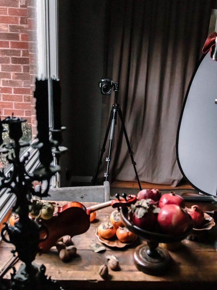 Example of chiaroscuro effect in food photography. A wooden table with fruit, black candle sticks and a violin and a large window showing directional light.