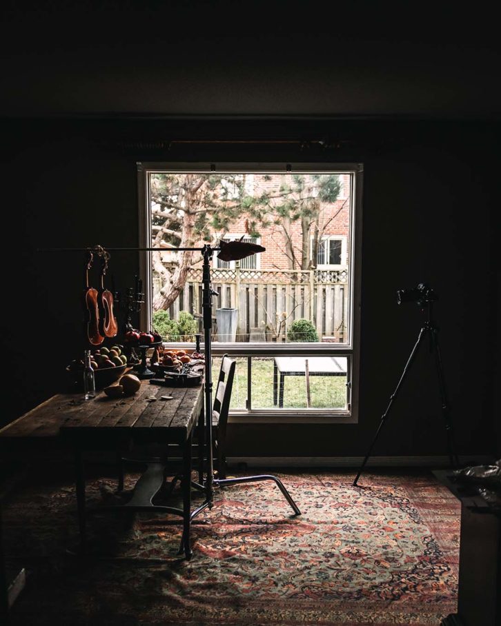 Example of chiaroscuro effect in food photography. A wooden table with fruit, black candle sticks and a violin and a large window showing directional light. Wide view.