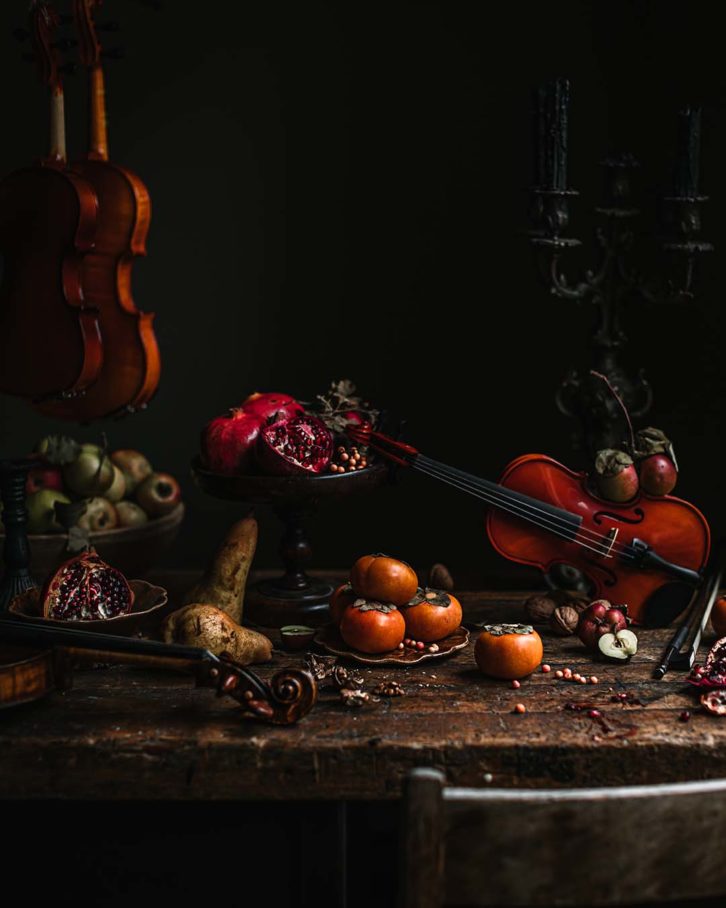 Example of chiaroscuro effect and natural light in food photography. A wooden table with fruit, black candle sticks and a violin. 