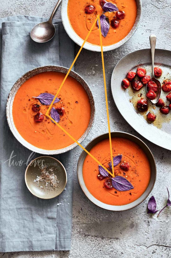 Grey marble background with three bowls filled with tomato soup and purple basil leaves ontop. 