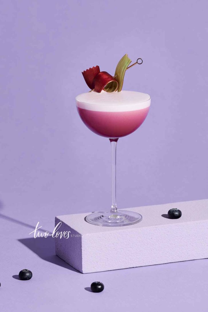 Bright purple background with a single tall stem cocktail glass filled with a berry foam cocktail.  