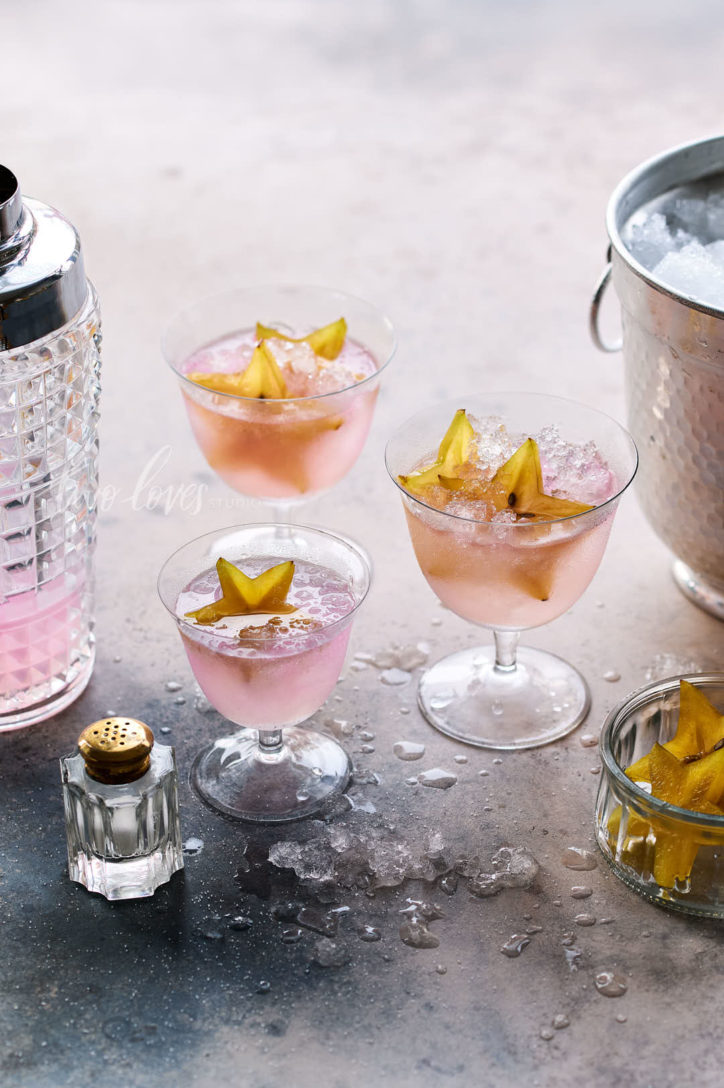 Marble background with three cocktail glasses filled with pink liquid and crushed ice. Star fruit and and ice bucket. 