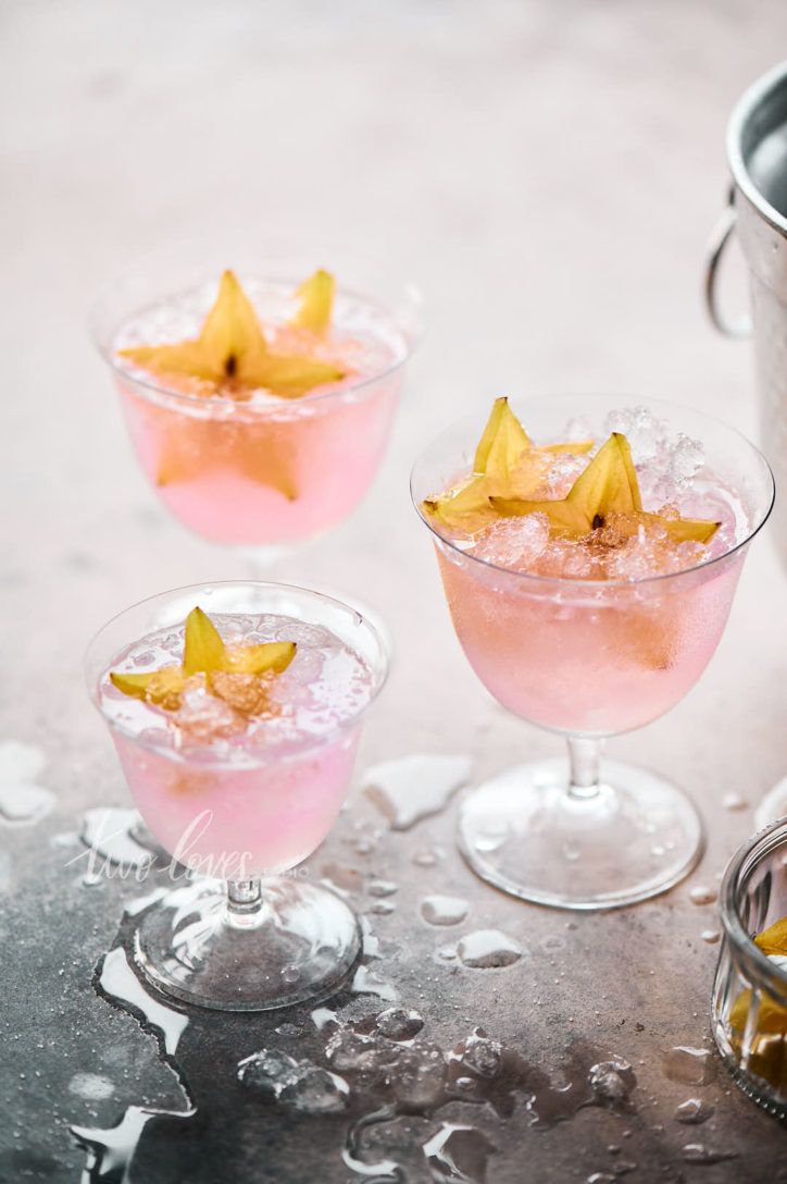 Marble background with three cocktail glasses filled with pink liquid and crushed ice. Decorated with a side of star fruit. 