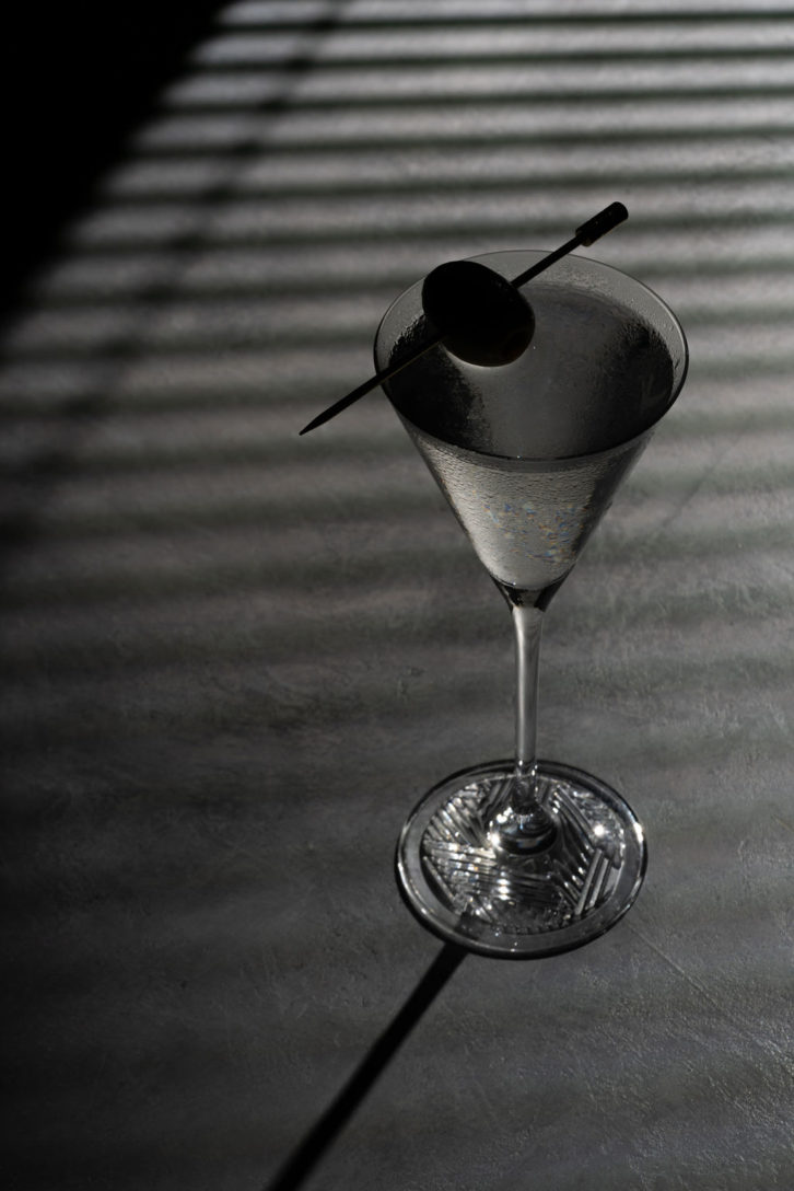 Dark background with a single martini glass on a dark background with an olive garnish. Too dark it's hard to see the image. 