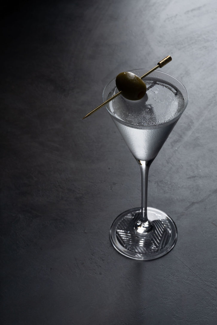 Dark grey background with a martini glass and a olive garnish. 