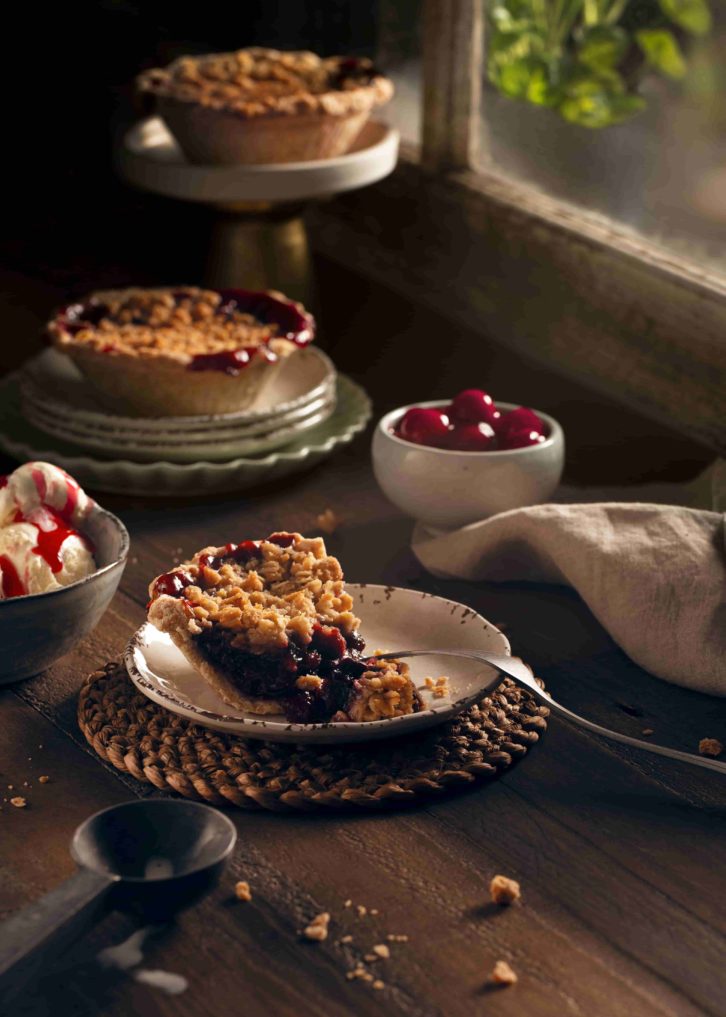 Wooden table with cheery pie pieces on while ceramic dishes. A bowl of ice cream as decoration with a lighted faux window in the background. 