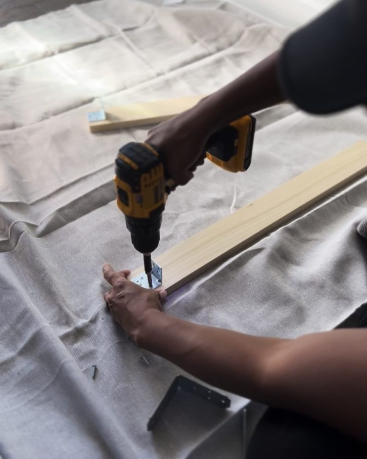 White table cloth on the ground, with two planks of wood and hands drilling in brackets. 