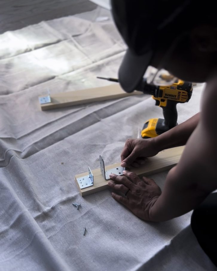 White table cloth on the ground, with two planks of wood, an electric screwdriver drilling in two bracket frames to hold the window. 