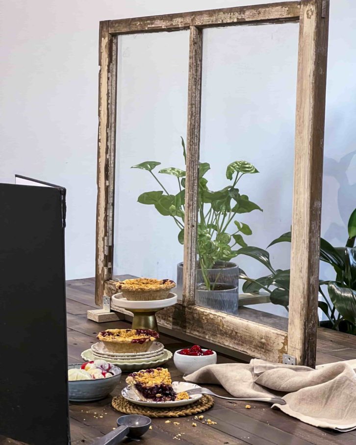 Wooden table decorated with faux plants being a faux window frame. Three cherry pies on white stands. A bowl of ice cream as decoration and a reflector board in the fore-ground. 