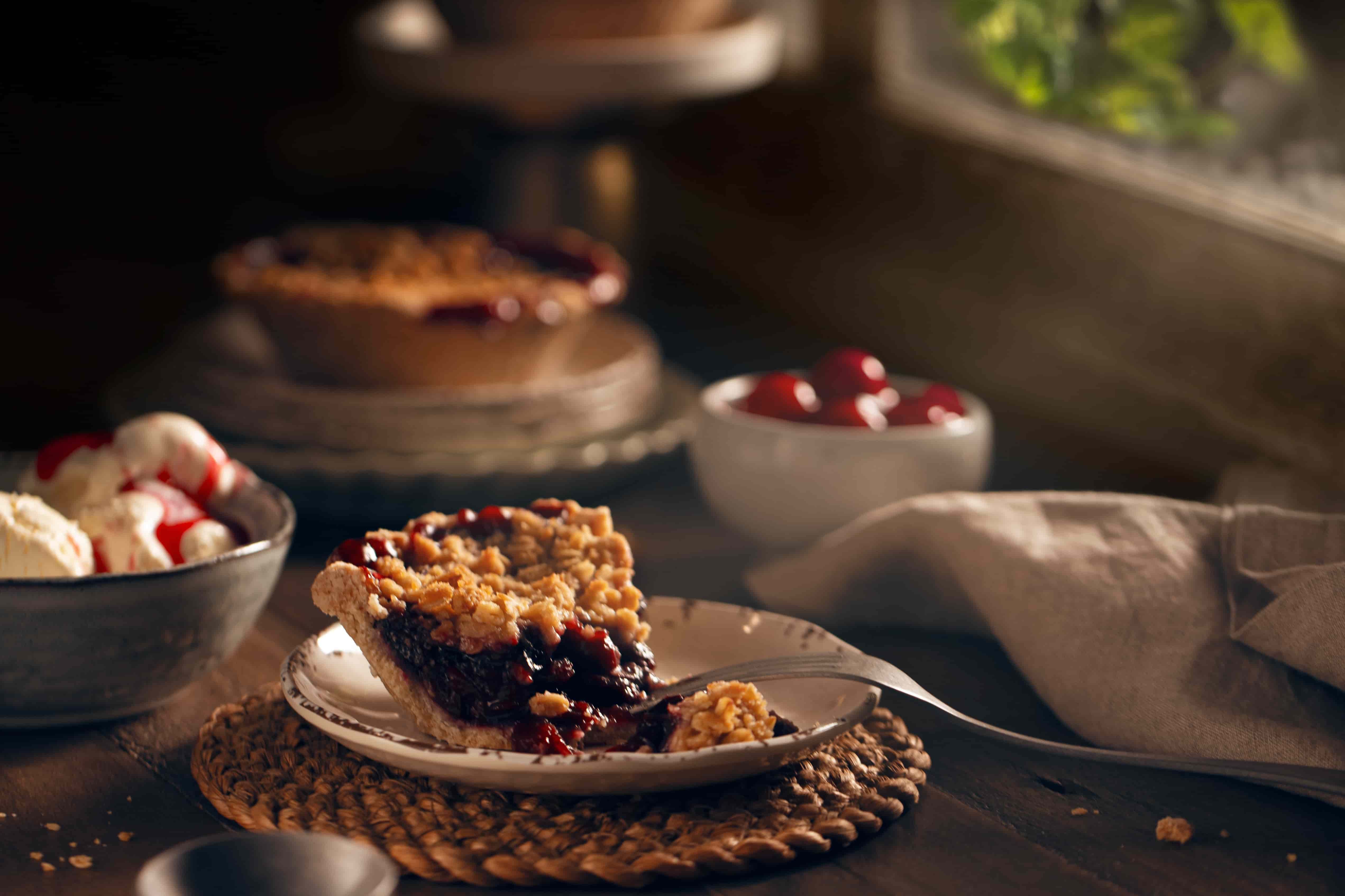 Wooden table with cheery pie pieces on while ceramic dishes. A bowl of ice cream as decoration with a lighted faux window in the background, showing a beam of light coming though, imitating sun light. 