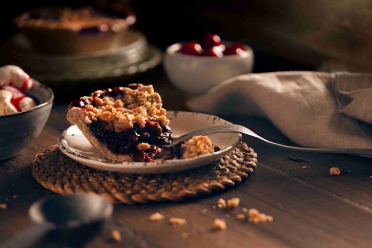 Wooden table with a single piece of cheery pie on while ceramic dish. A bowl of ice cream to the left as decoration with a lighted faux window in the background. 