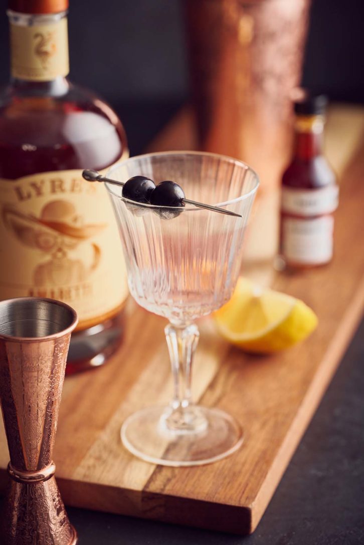 Shot of a glass with a steam and cherry garnishes, on a wooden chopping board. Showing how to avoid reflections in photography by using textured glass wear. 