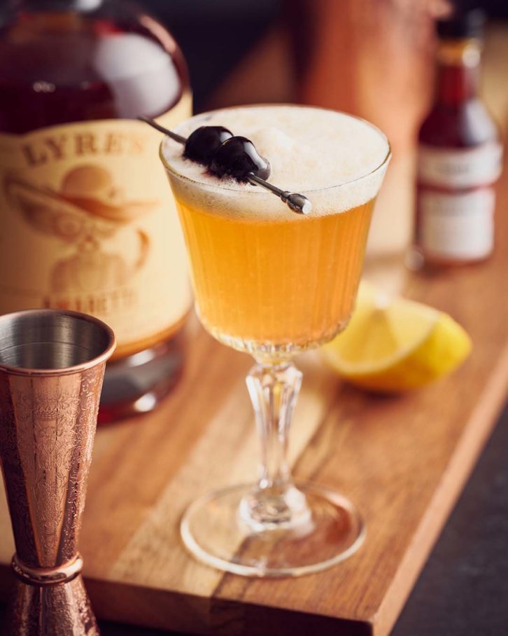 Hero virgin amaretto sour with a cherry garnish in a glass with a steam, on a wooden chopping board. 