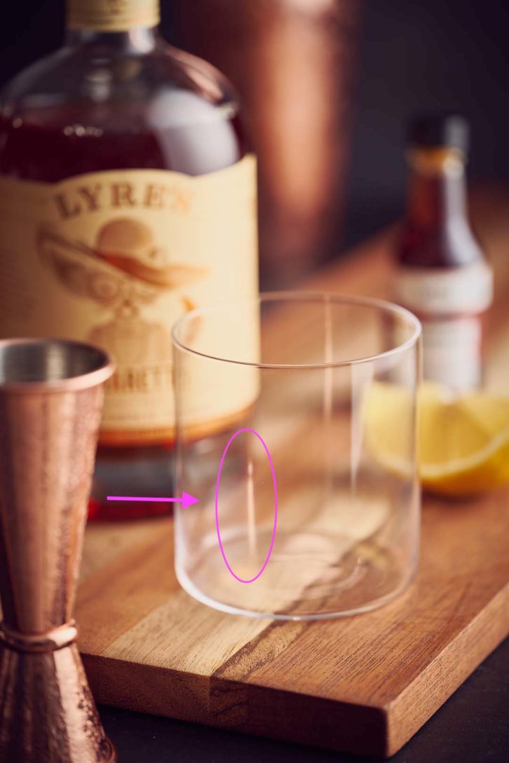 Thin walled clear glass, on a wooden chopping board, showing the reflection of the jigger. 