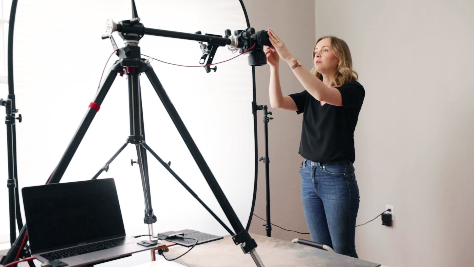 A person setting up a camera on a tripod, which is tethered to a computer, with lighting equipment in the background