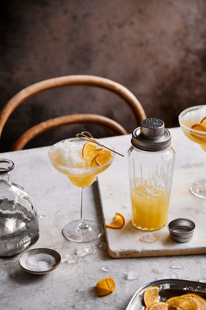 Marble table with wooden chair, glass cocktail shaker and cocktail glass filled with an orange crushed ice cocktail.