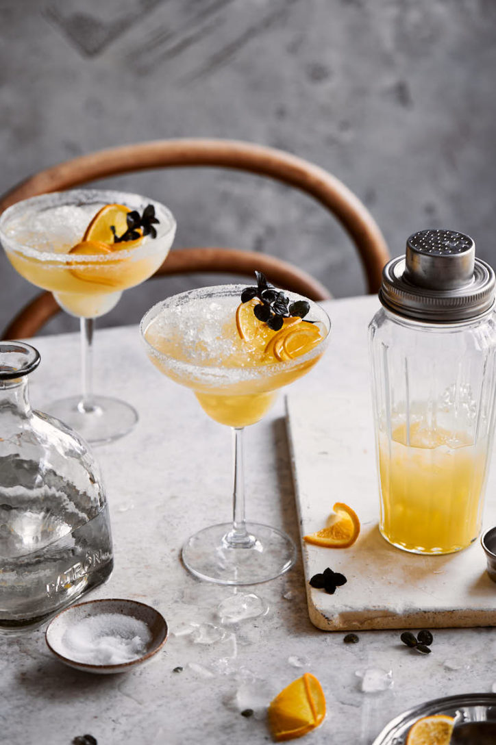 Marble table with wooden chair, glass cocktail shaker and cocktail glass filled with an orange crushed ice cocktail.