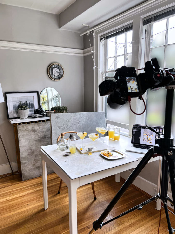 Wide angle set up shot showing a a large white table with a marble background. Three cocktail glasses filled with an orange tequila mix.