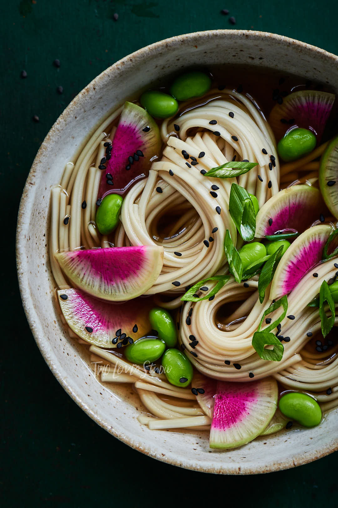 Dark green background with a speckled large bowl, with noodles and slices of radishes. Sprinkled with black sesame seeds and edamame. 