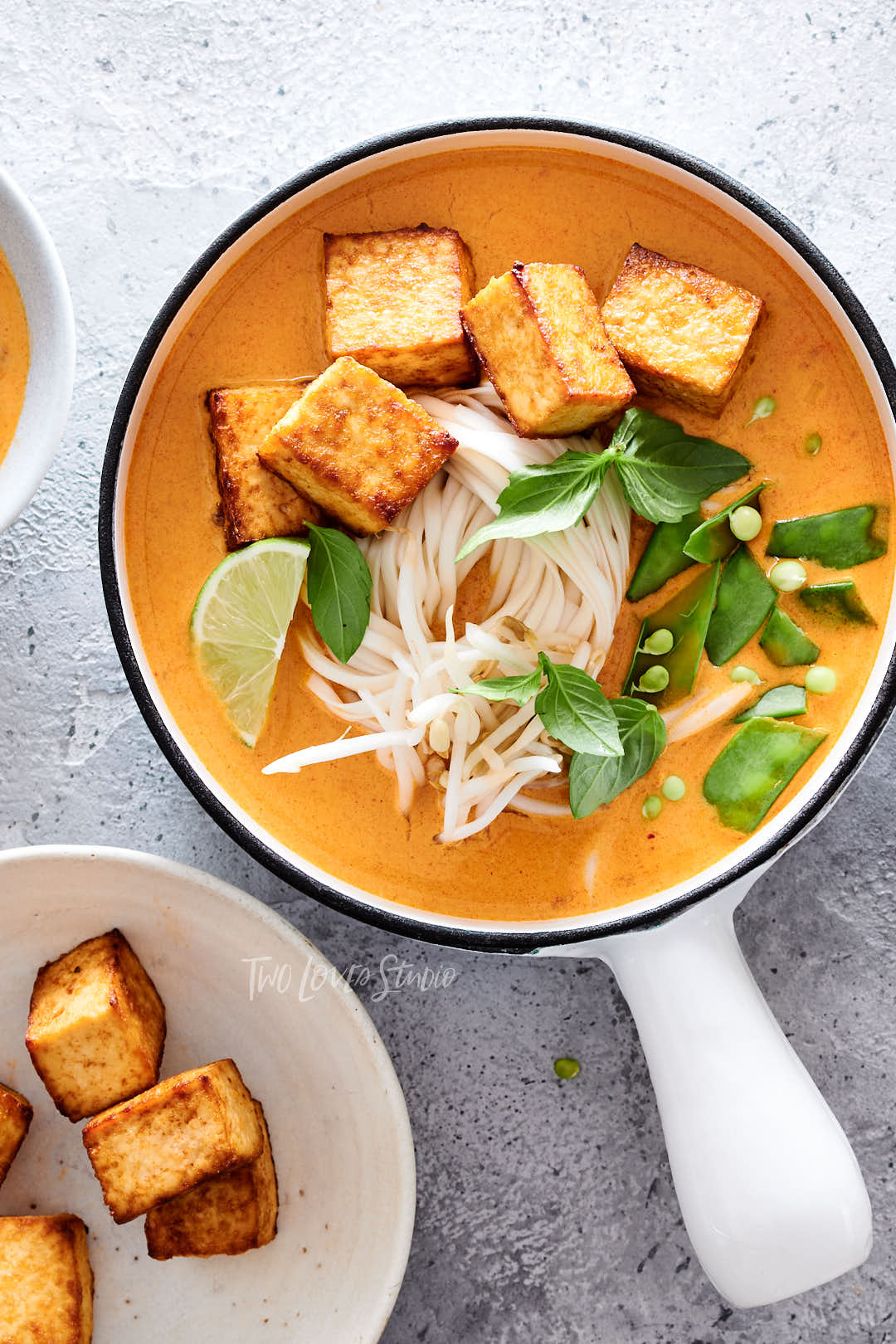 Grey background, with a white handled bowl. Orange broth with tofu. 
