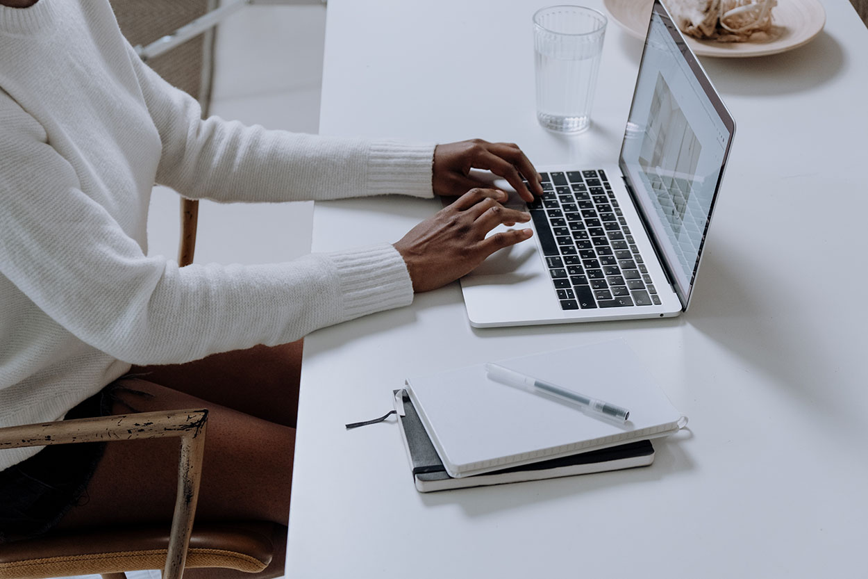 person typing on a laptop with a note pad, sitting at a desk.