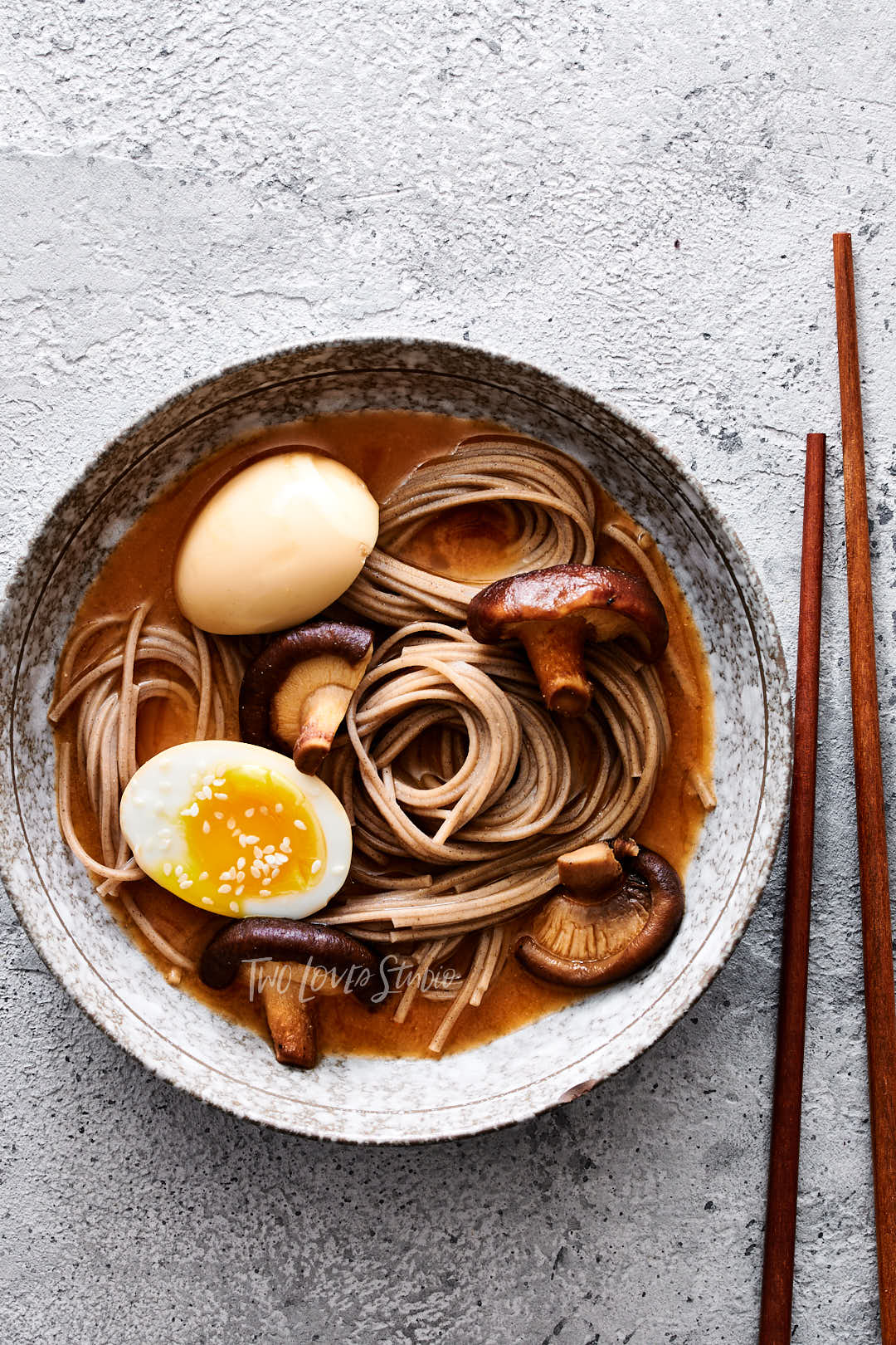 Grey background with a speckled bowl, noodle swirls with a dark broth, dark mushrooms and a boiled egg. 