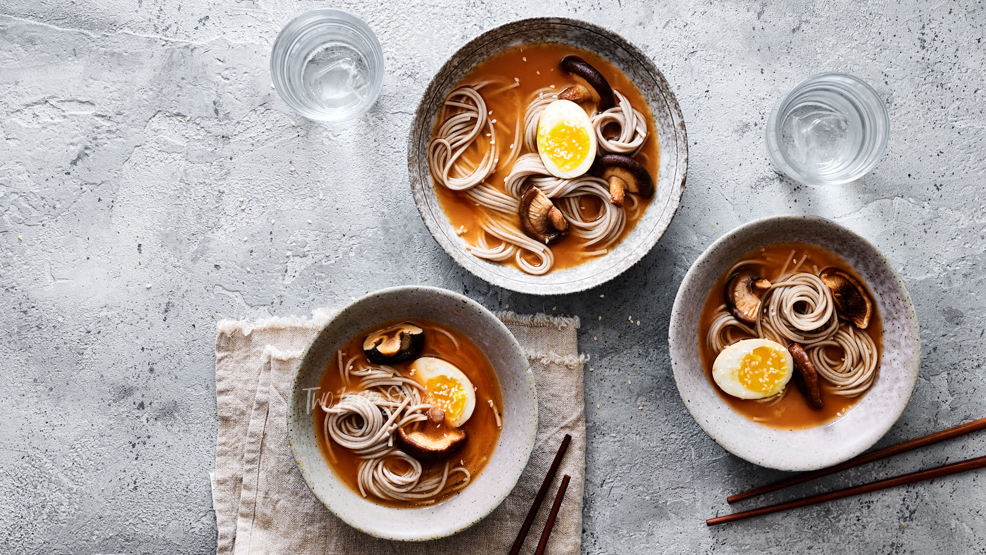 Three bowls filled with a dark broth, noodles, mushrooms and boiled eggs.
