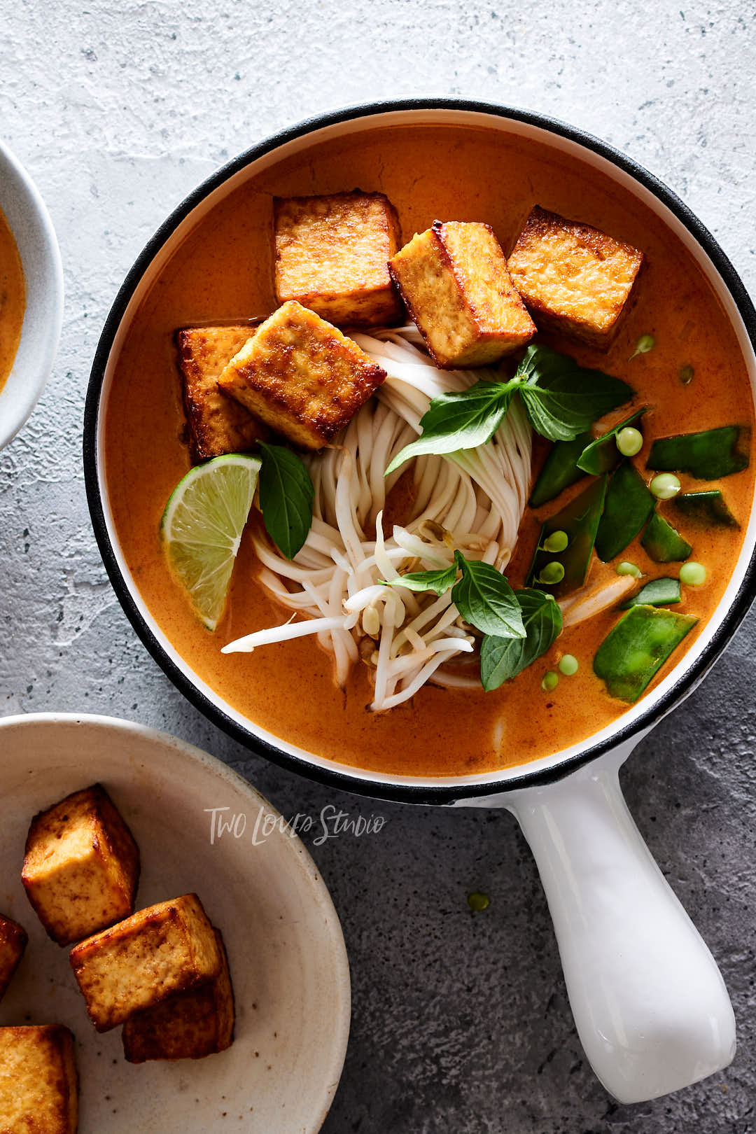 Grey background, with a white handled bowl. Orange broth with tofu. 