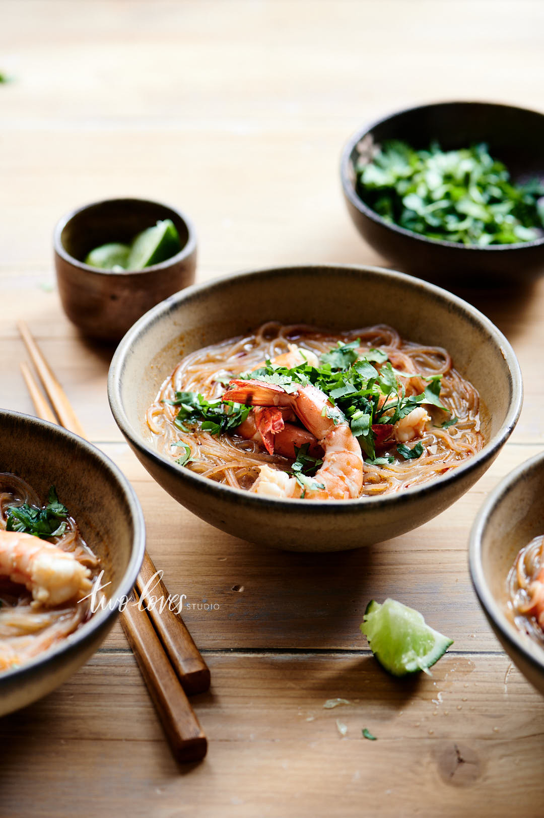 Wooden table with a pottery bowl filled with shoe string noodles, prawns and a sprinkling with green garnish.