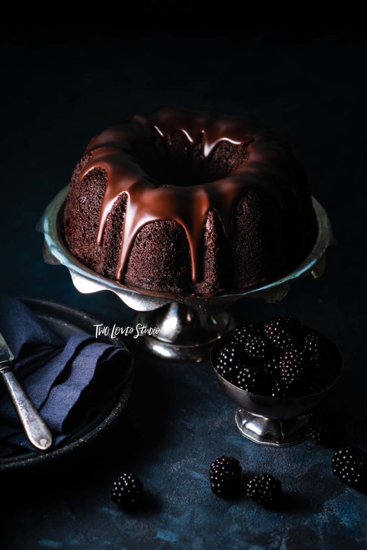 Dark blue background with blackberries and a silver cake tray and a chocolate bunt cake with chocolate dripped icing.
