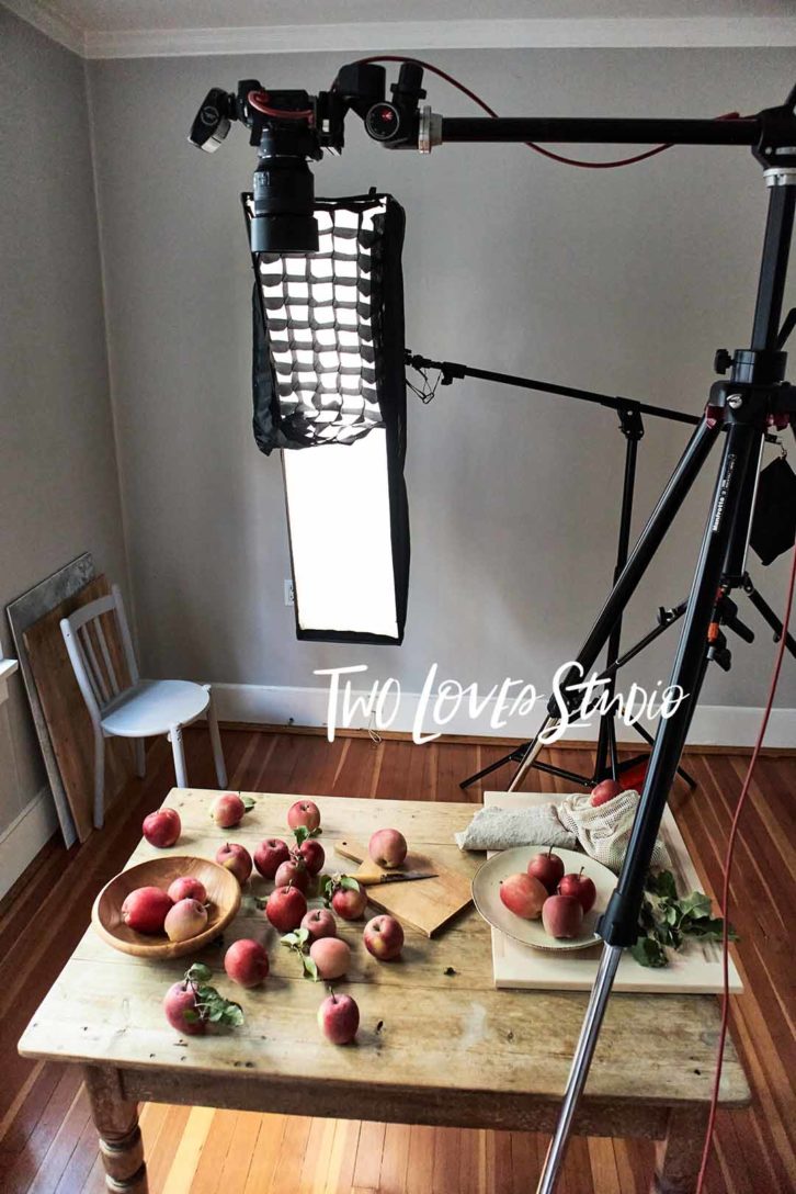 Wooden table with red apples and a behind the scenes shot of strip box set up in a studio for modifiers for food photography.