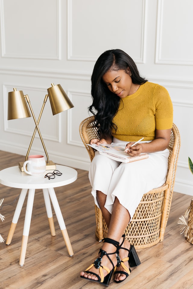 Women sitting in a wicker chair next to a small white side table working on a notebook