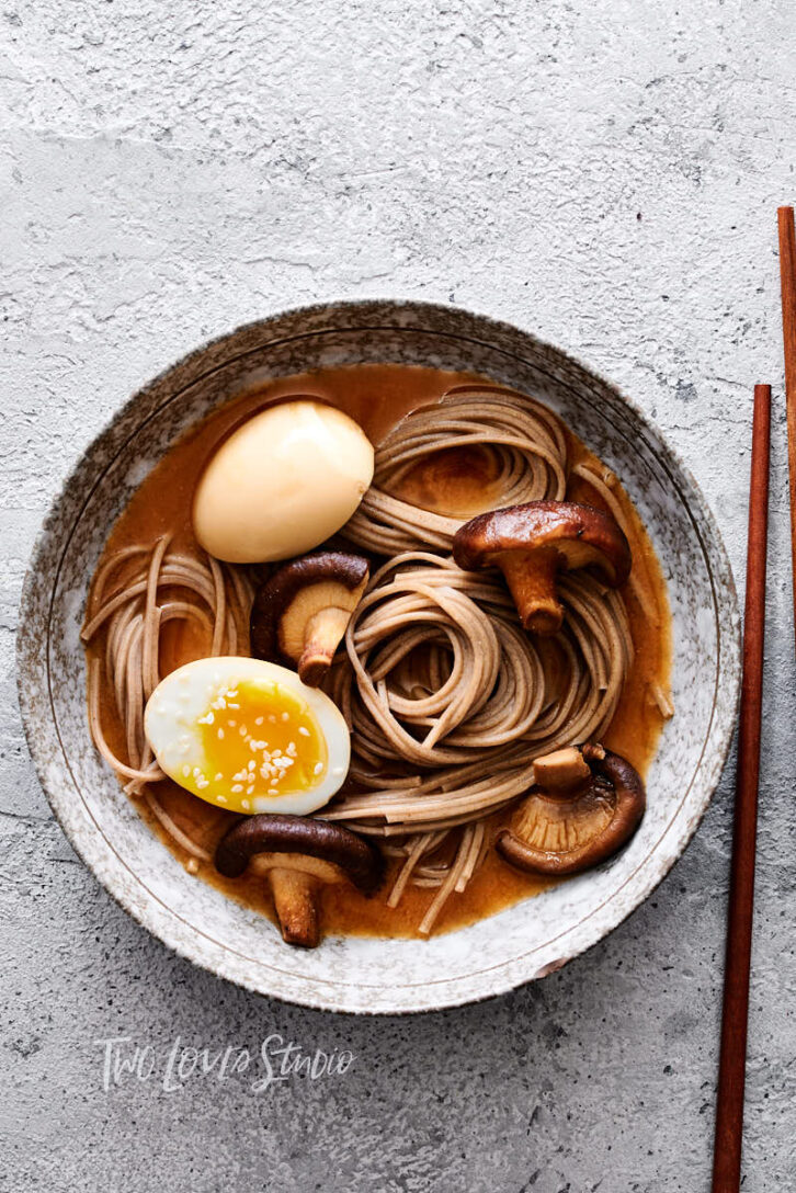 Grey stone background with a Japanese marble bowl with brown soup, mushrooms, noodles and a boiled egg.  