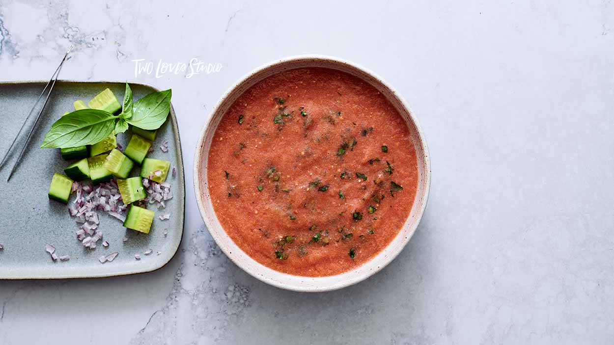 How-to-style soup, gazpacho. A bowl of red soup on a marble background with a plate of garnishes and toppings.