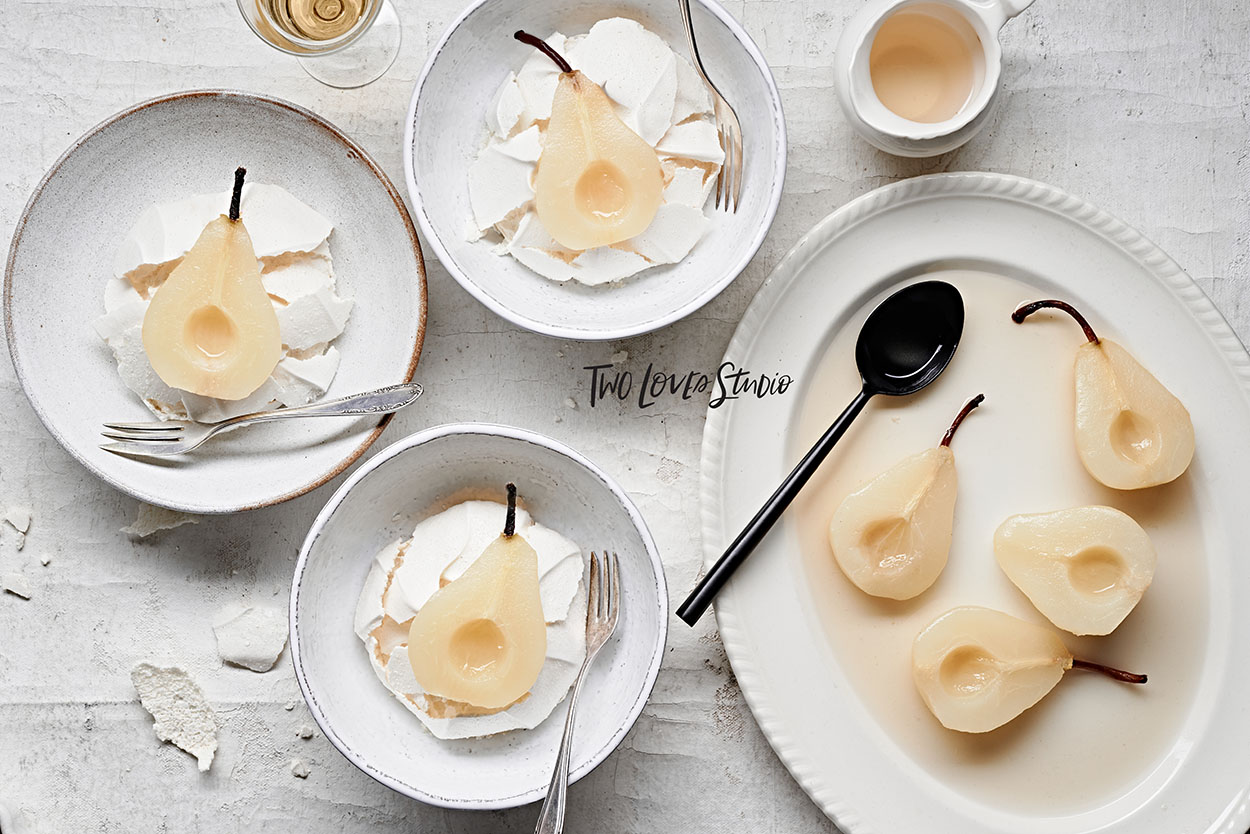 White wine poached pears with meringue nests on a white background with white plates and a large black spoon.