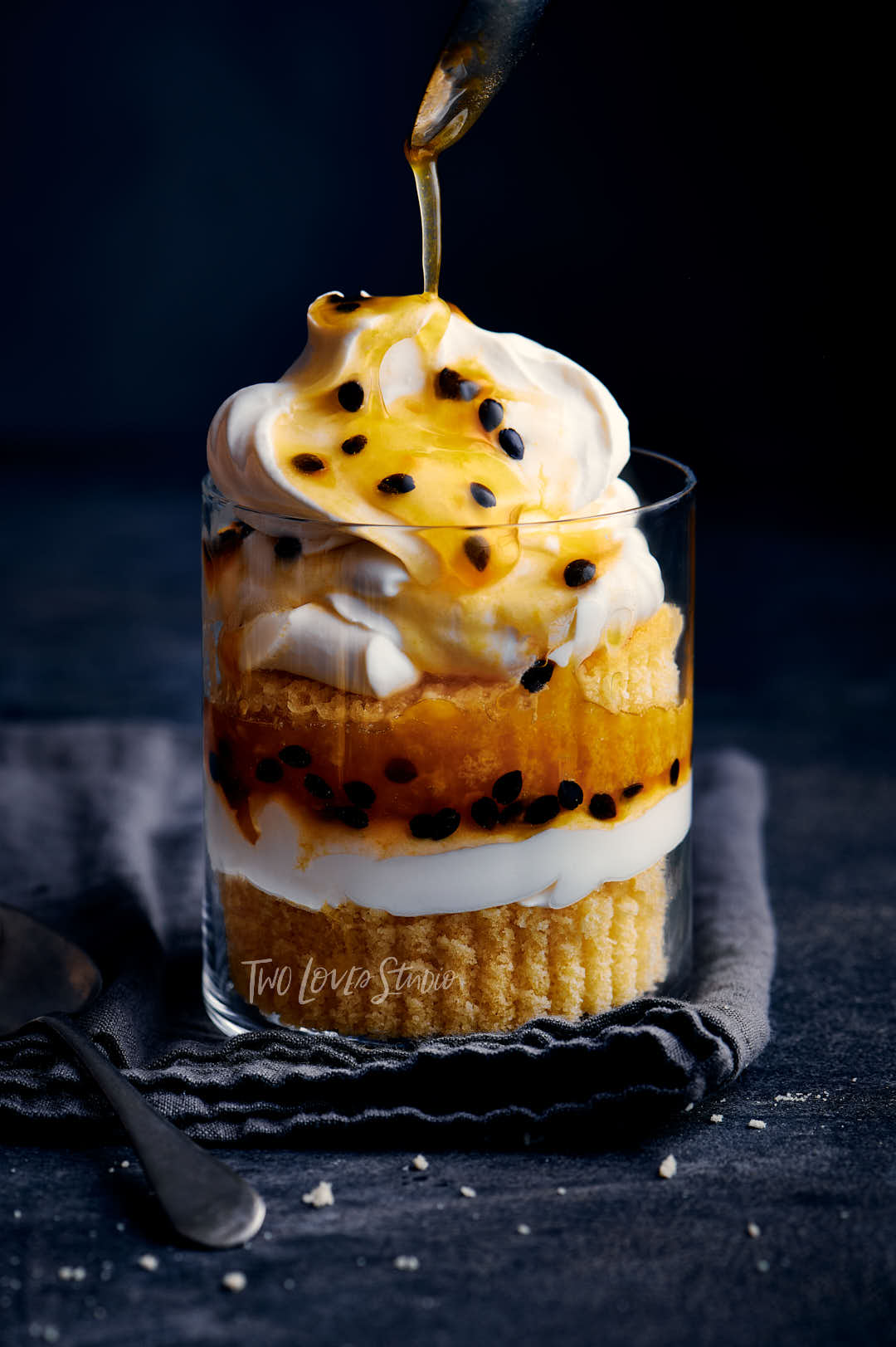 Passionfruit trifle with layers of cake and cream on a dark blue background. A drizzle from a spoon is coming from the top of the image.