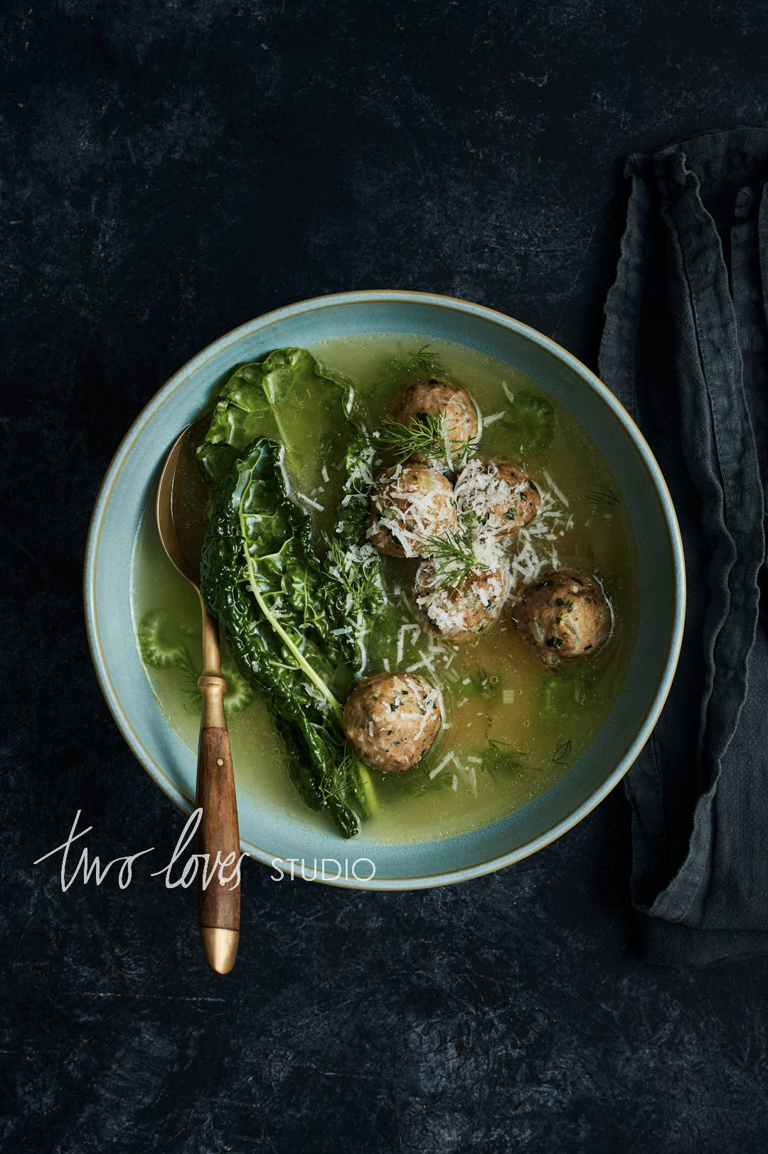 Chicken meatball soup with large kale leaves. A wooden brass spoon into the bowl with a dark linen.