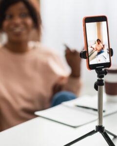 A person sits on a couch, facing a smartphone mounted on a tripod. The phone is recording with a notepad.