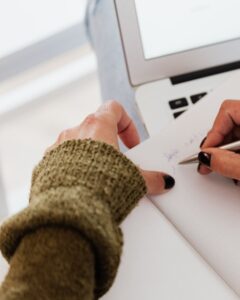 A person wearing a green knit sweater is writing in a notebook with a pen. An open laptop is visible in the background.